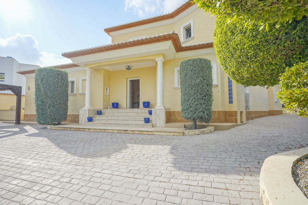 Entrada principal con porche de columnas blancas, escalinata de piedra decorada con macetas azules y amplio camino pavimentado rodeado de setos ornamentales.