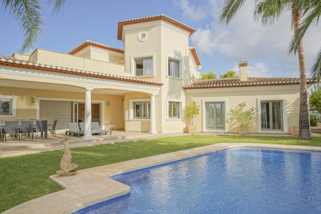 Fachada de la villa con piscina privada, césped y porche cubierto con columnas. Destaca la arquitectura tradicional con techos de teja y carpintería blanca.