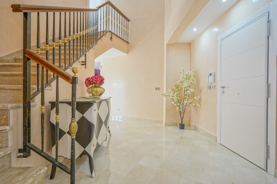 Hall de entrada con suelos de mármol pulido, una escalera de piedra con barandilla de hierro forjado y una cómoda decorativa con patrón de rombos.