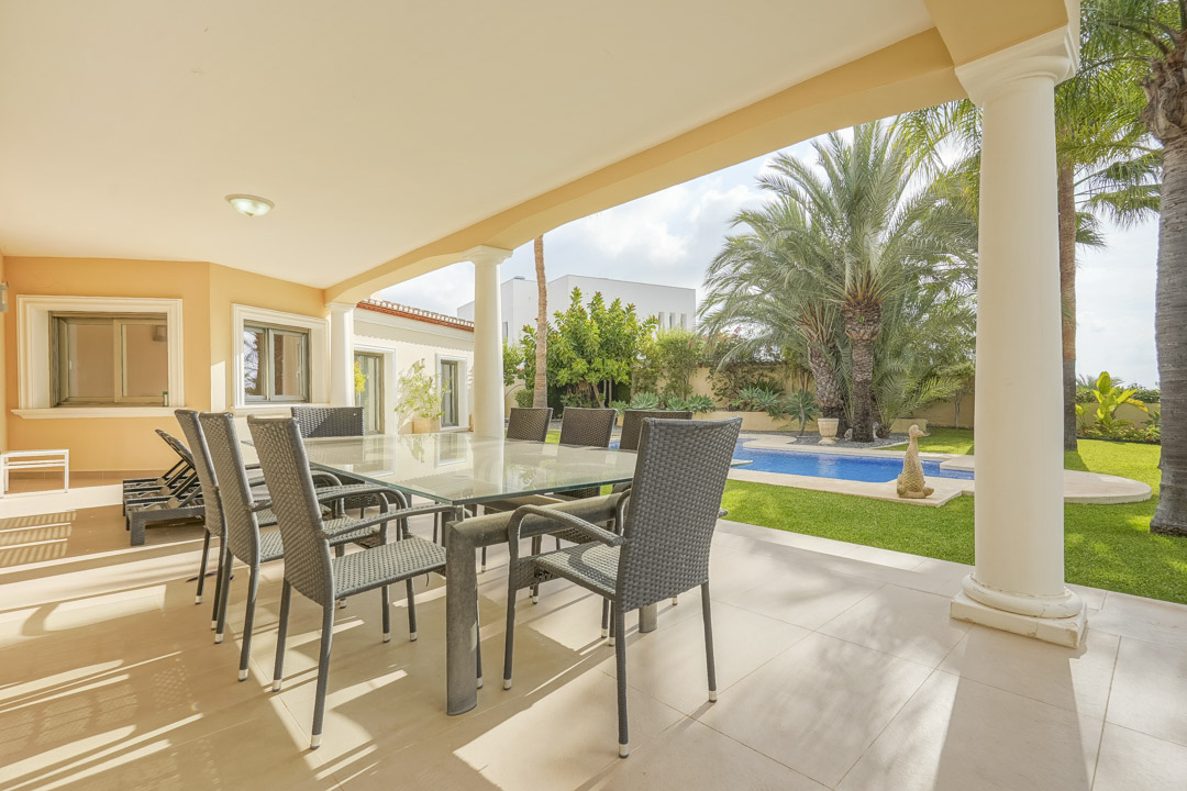 Terraza cubierta con columnas blancas, comedor exterior con mesa de cristal y vistas directas a la piscina y al jardín mediterráneo.