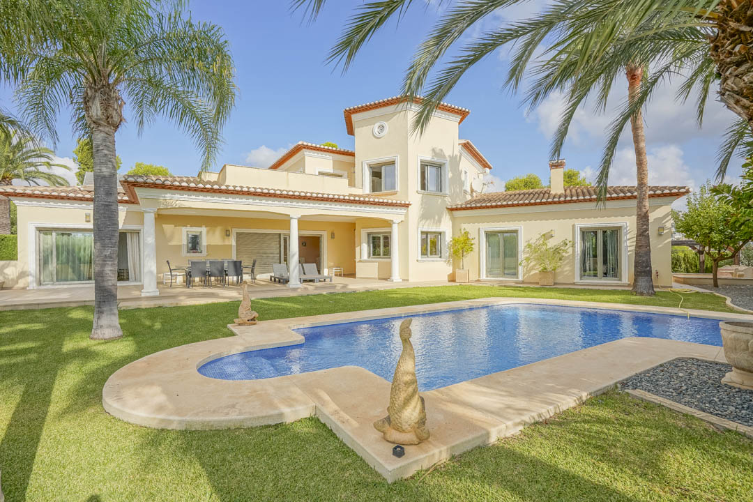 Vista exterior de la villa con piscina privada, jardín de césped con palmeras y porche cubierto sostenido por columnas blancas bajo una arquitectura tradicional.