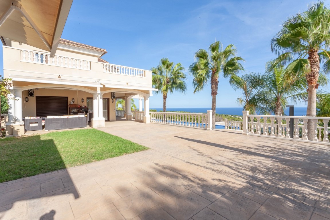 Amplia terraza exterior con suelo de piedra, balaustrada clásica blanca y vistas panorámicas al mar. Incluye un porche cubierto con mobiliario y palmeras.