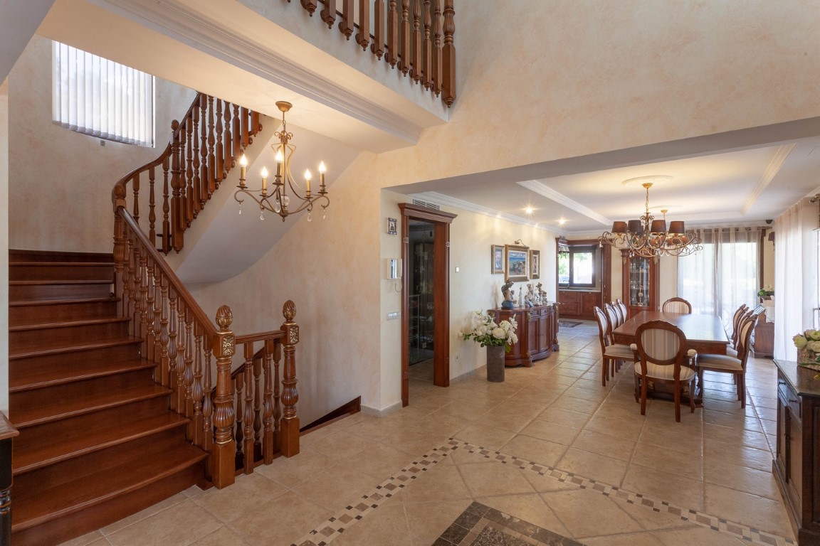 Amplio vestíbulo con escalera de madera tallada y suelos cerámicos decorados que conectan con un comedor formal de estilo clásico y techos altos.