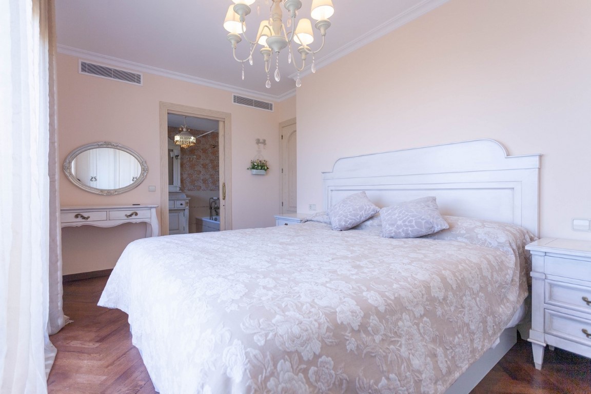 Dormitorio con cabecero de madera blanca, suelos de parqué y acceso a un baño en suite con bañera y tocador de estilo clásico.