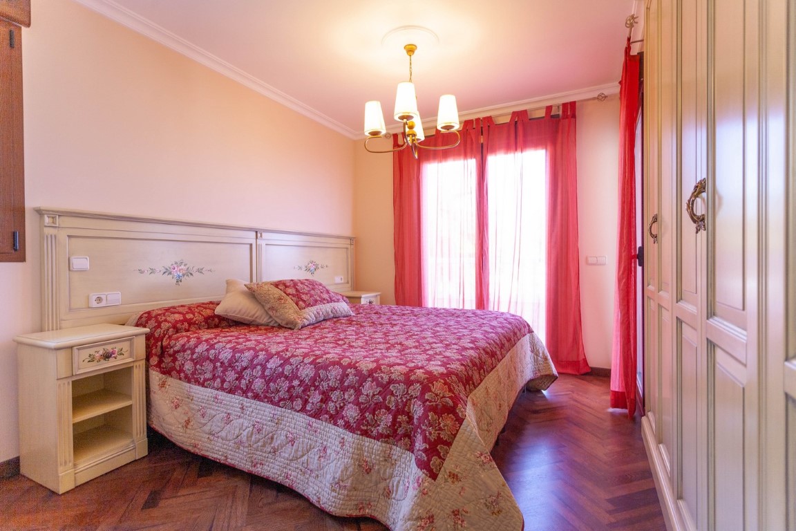 Dormitorio con suelo de parqué en espiga, armario empotrado color crema y acceso al balcón. Incluye mobiliario clásico con detalles florales y cortinas rojas.