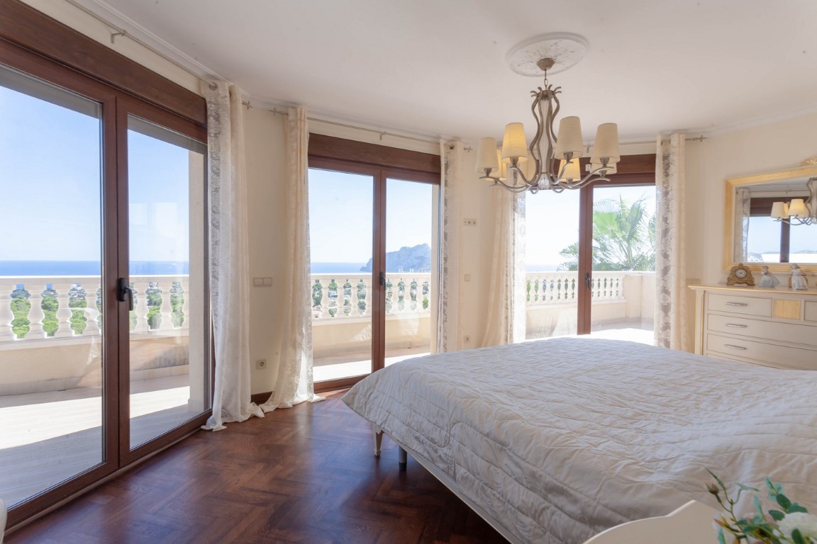 Dormitorio con suelos de parqué, lámpara de araña clásica y amplios ventanales que ofrecen vistas al mar y acceso directo al balcón con balaustrada.