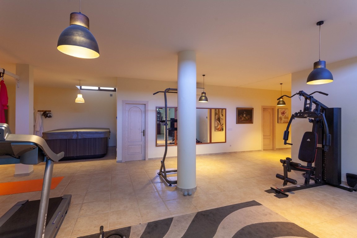 Gimnasio privado y zona de bienestar con máquinas de entrenamiento, jacuzzi cubierto y suelos de gres, iluminado por lámparas colgantes de estilo industrial.