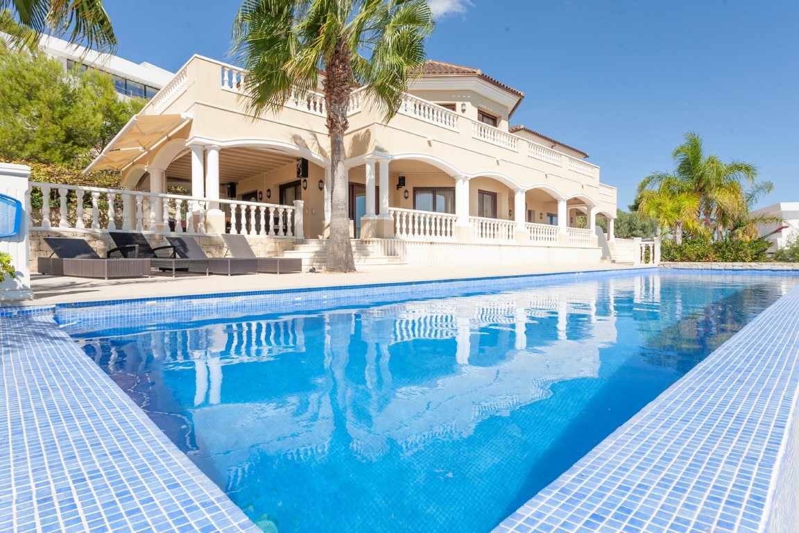 Piscina de mosaico azul frente a una villa de estilo clásico con arcos, balaustradas blancas y amplias terrazas cubiertas rodeadas de palmeras.