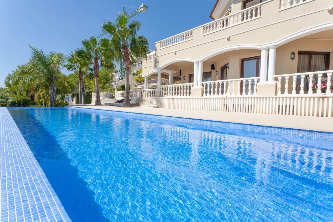 Piscina de mosaico azul frente a una villa de estilo mediterráneo con terrazas porticadas, balaustradas blancas y palmeras bajo un cielo despejado.