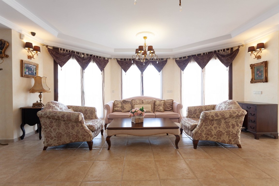 Salón luminoso con suelos de gres, mobiliario de estilo clásico, sillones florales y amplios ventanales que aportan abundante luz natural.