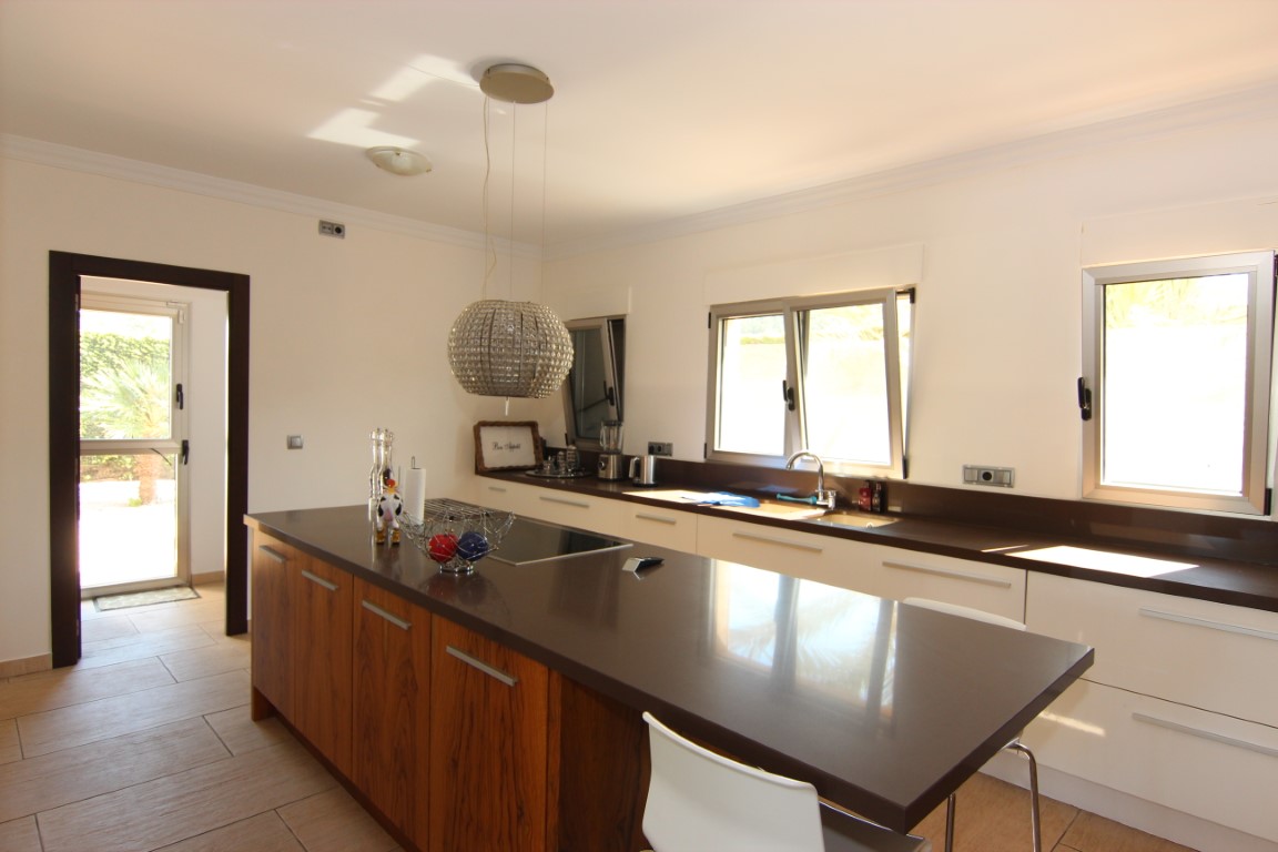 Cocina espaciosa con isla central de madera, encimeras de cuarzo oscuro y lámpara de cristal. Dispone de ventanales de aluminio y acceso directo al jardín.