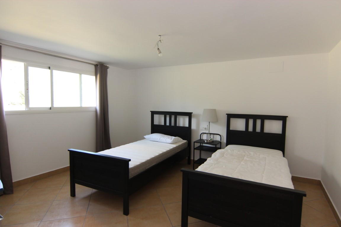 Dormitorio con dos camas individuales de madera oscura, suelos de gres cerámico y luz natural a través de una amplia ventana corredera con cortinas.