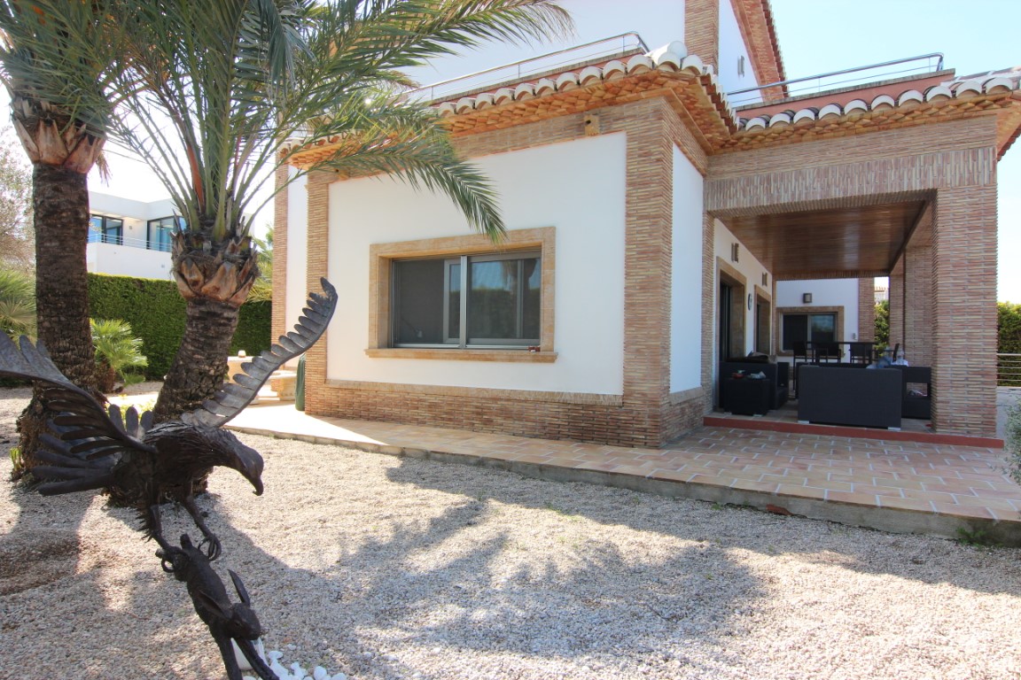 Exterior de la villa con fachada de ladrillo visto, porche cubierto con mobiliario de exterior y jardín de grava con palmeras y escultura decorativa.