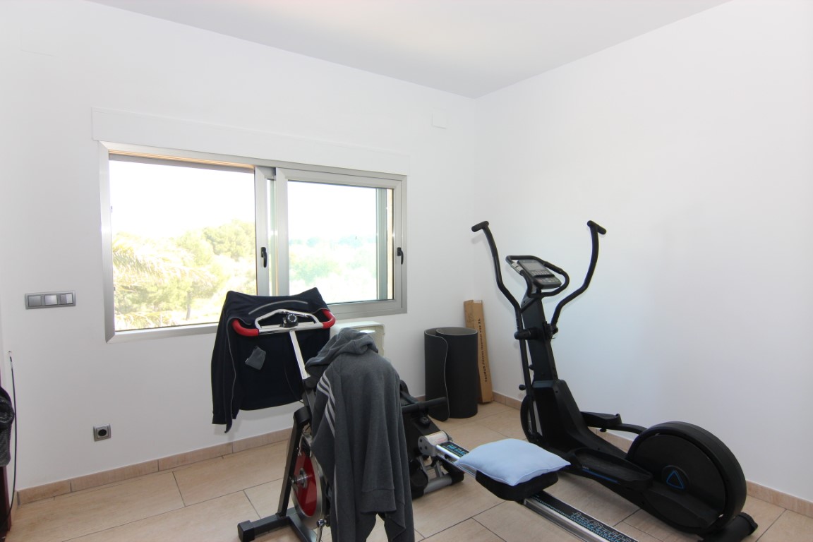 Gimnasio privado equipado con máquinas de fitness, suelos cerámicos y un gran ventanal que aporta abundante luz natural y vistas al entorno.
