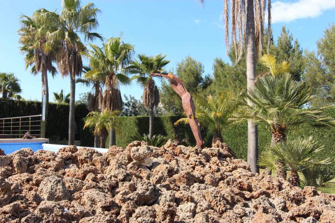 Jardín mediterráneo con palmeras, una escultura abstracta de metal y rocas decorativas junto a la zona de la piscina.