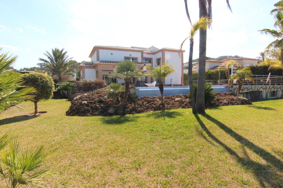 Vista exterior de la villa con jardín mediterráneo, césped extenso y piscina elevada sobre rocalla natural. La fachada combina muros blancos con detalles de ladrillo.