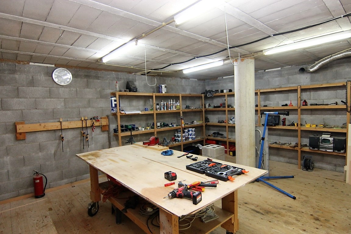 Amplio taller en el sótano con paredes de bloques de hormigón, estanterías de madera perimetrales y un gran banco de trabajo central sobre suelo de madera.