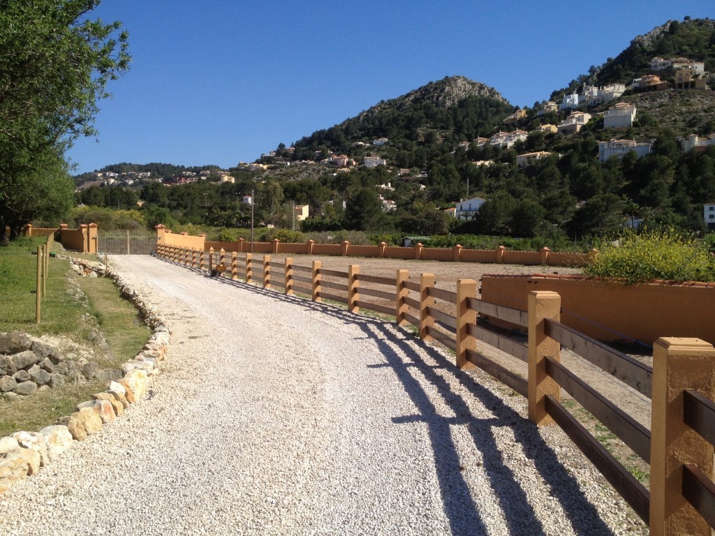 Camino de entrada de grava blanca flanqueado por una valla de madera y muros perimetrales, con vistas despejadas a las colinas y vegetación local.