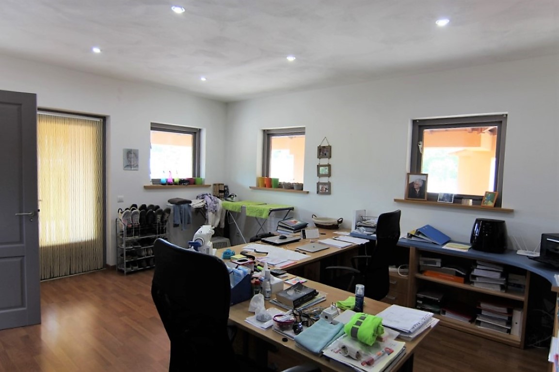 Despacho funcional con suelo de madera, iluminación empotrada y múltiples ventanas. El espacio cuenta con una amplia mesa de trabajo en L y estanterías integradas.