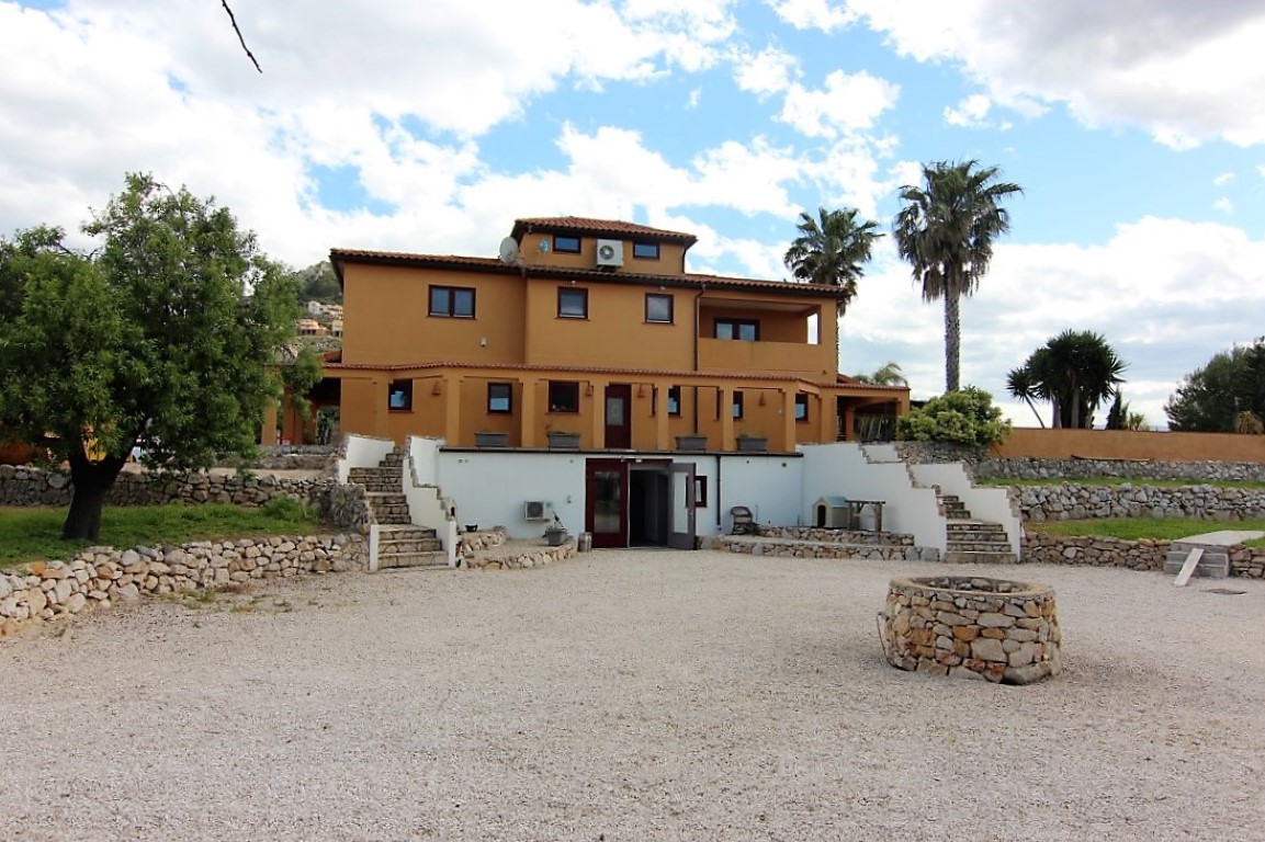 Vista exterior de la finca de tres niveles con fachada ocre, muros de piedra seca y un amplio patio de grava que incluye un pozo decorativo.