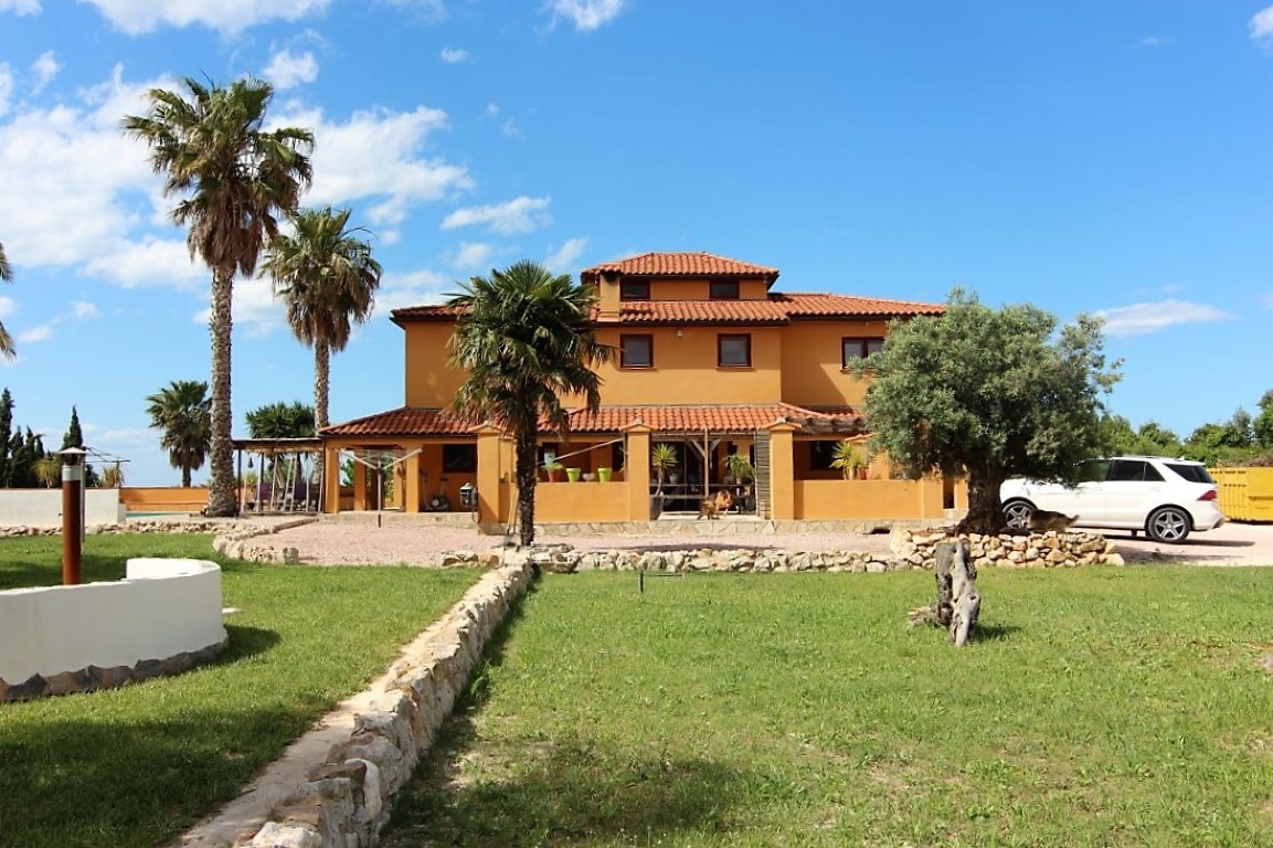 Vista exterior de la finca de tres niveles con fachada ocre, techos de teja y amplios jardines con palmeras y muros de piedra natural.