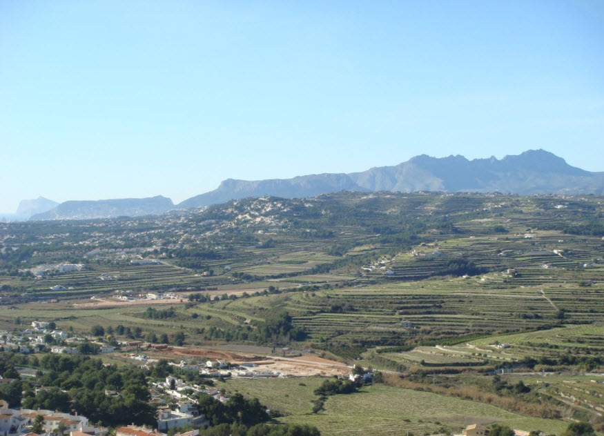 Vista panorámica de los bancales agrícolas y el relieve montañoso de Benitachell bajo un cielo despejado, mostrando el entorno natural del proyecto.