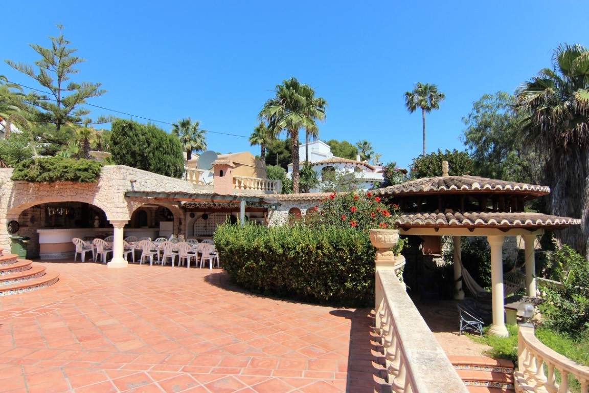 Amplia terraza de baldosas de terracota con zona de comedor cubierta bajo arcos de piedra y un cenador rústico rodeado de vegetación mediterránea y palmeras.
