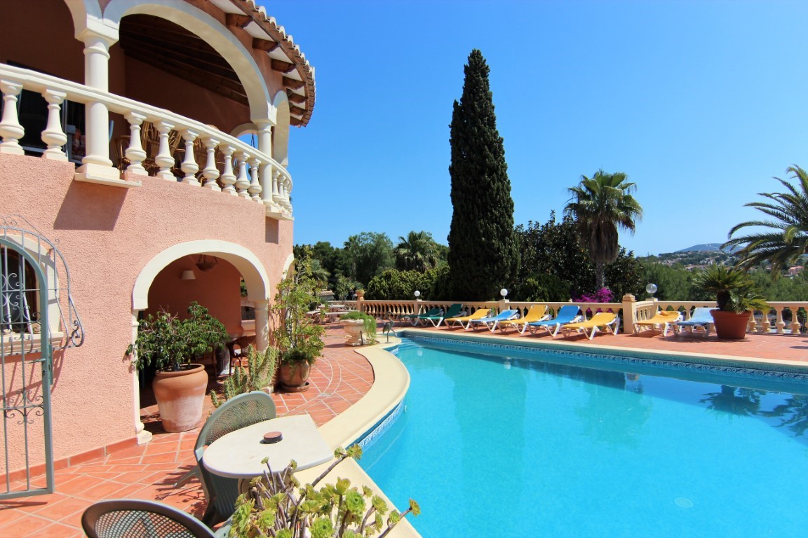 Amplia terraza de terracota con piscina de forma curva, rodeada de tumbonas y vegetación mediterránea bajo una fachada con arcos y balaustradas blancas.