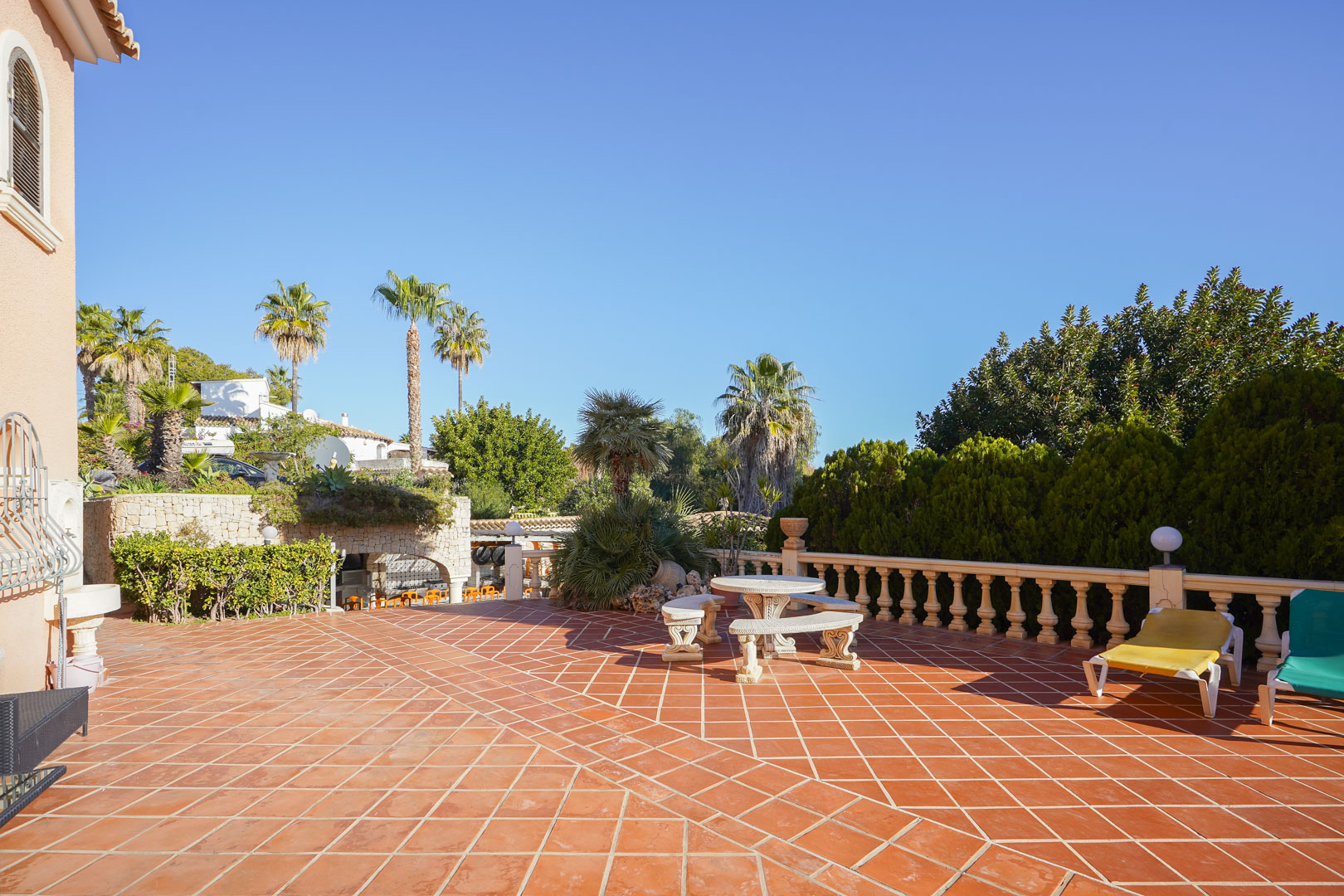 Amplia terraza exterior con baldosas de terracota, balaustrada de piedra y mobiliario de jardín, rodeada de palmeras y vegetación mediterránea bajo un cielo despejado.