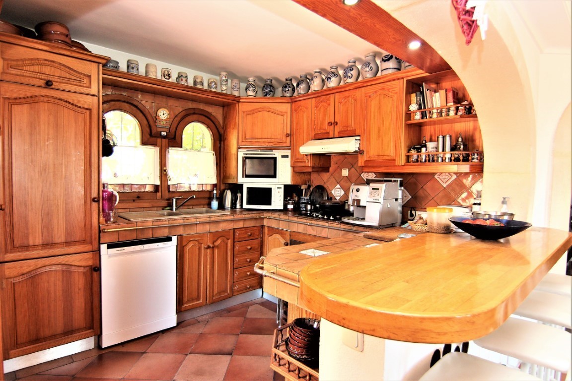 Cocina rústica con armarios de madera, encimeras de azulejo y barra de desayuno. Incluye electrodomésticos integrados, suelo de terracota y ventanas de arco tradicionales.