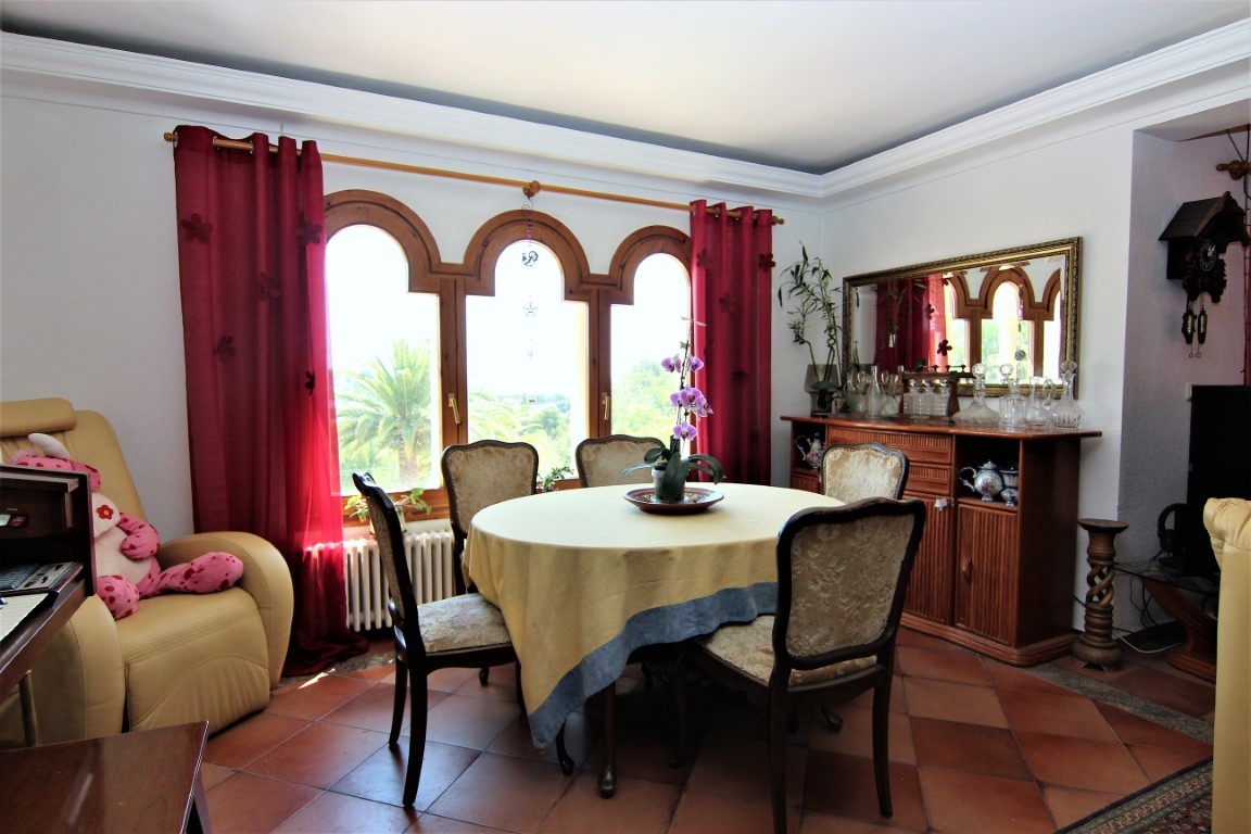 Comedor con mesa redonda, sillas clásicas y suelos de terracota. Destaca el ventanal de madera con tres arcos y vistas al jardín mediterráneo.