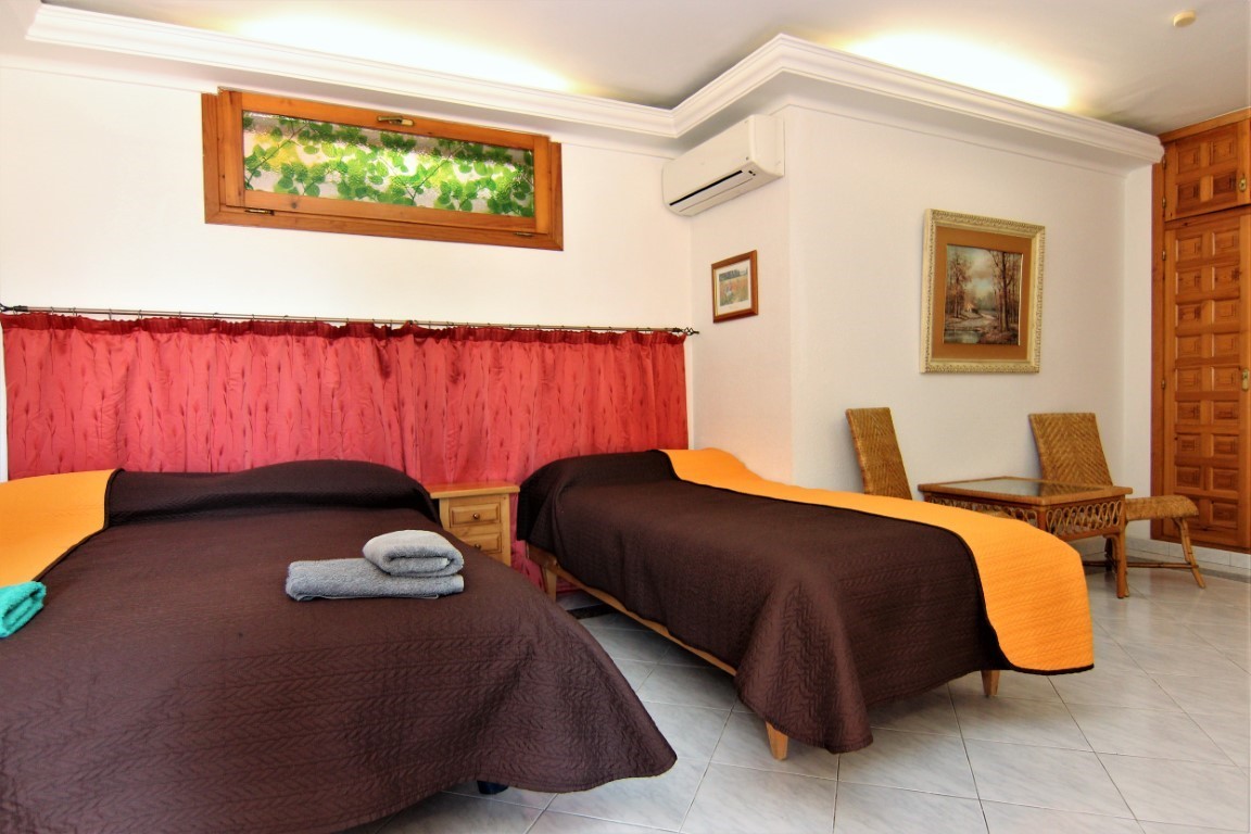 Dormitorio con dos camas, suelos cerámicos y aire acondicionado. Incluye mobiliario de mimbre, armario empotrado de madera y una ventana alta con marco de madera.