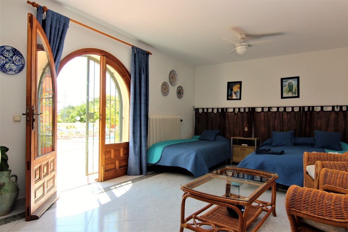 Dormitorio en planta baja con acceso directo a la terraza de la piscina mediante una puerta de madera en arco, muebles de mimbre y ventilador de techo.