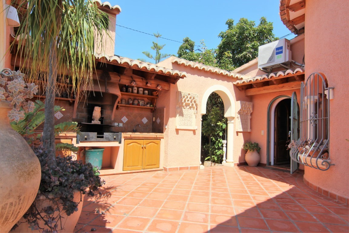 Patio exterior con cocina de verano techada, barbacoa integrada y suelos de terracota. Presenta un arco de estilo mediterráneo y acabados en tonos tierra.
