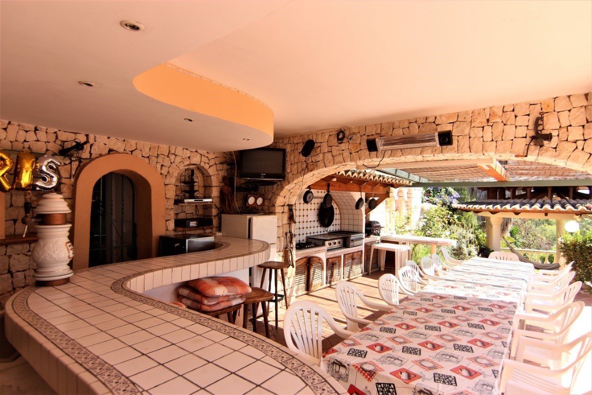 Terraza cubierta con bar de azulejos curvos, paredes de piedra natural y una amplia zona de comedor exterior equipada con cocina profesional.