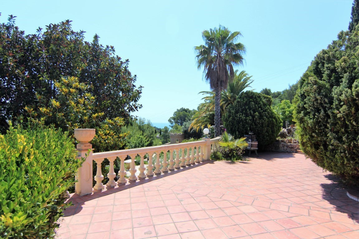 Terraza exterior con suelo de baldosas cerámicas y balaustrada clásica de piedra, rodeada de vegetación mediterránea y vistas lejanas al mar.