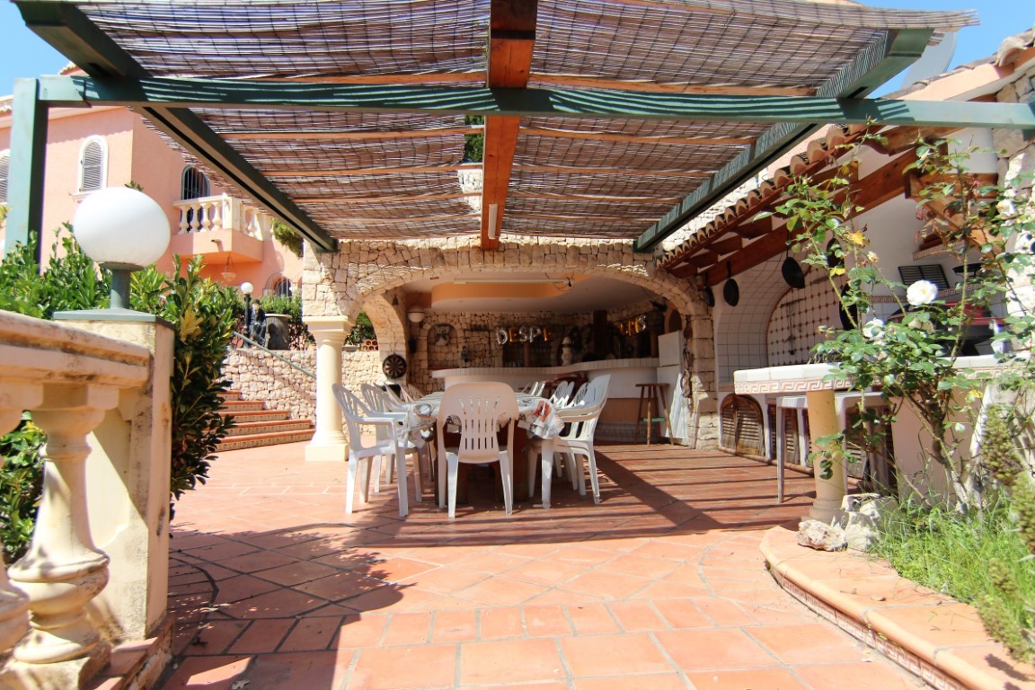 Terraza exterior cubierta con pérgola de cañizo, amplia zona de comedor y cocina de verano integrada bajo arcos de piedra natural y suelos de terracota.