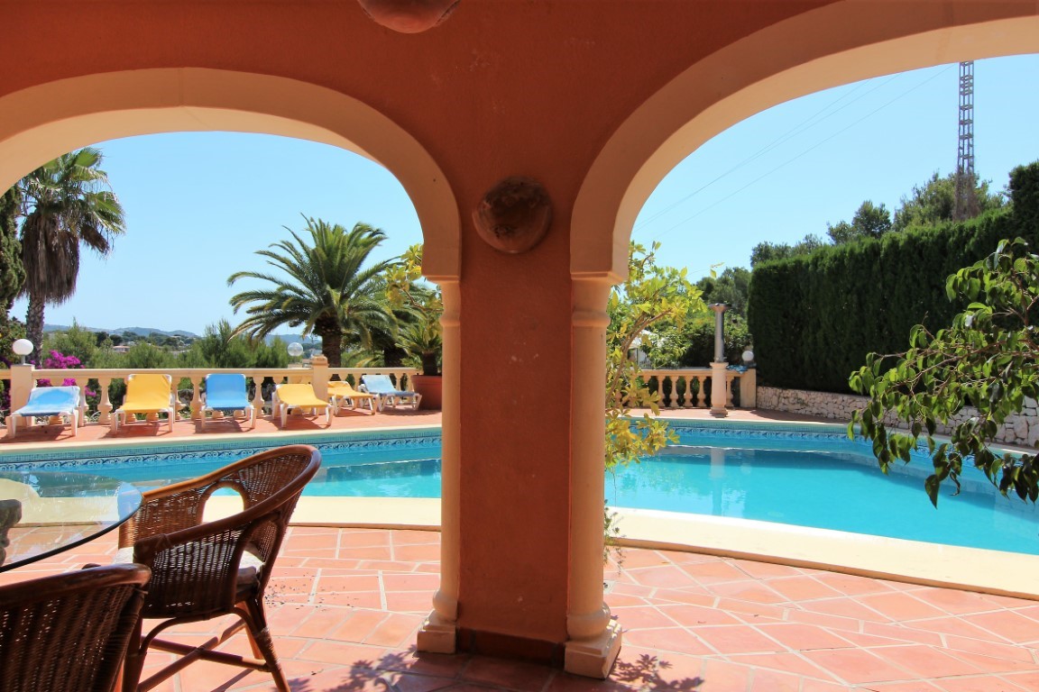 Vista desde el porche cubierto con arcos hacia la amplia piscina exterior, rodeada de suelos de terracota, tumbonas y vegetación mediterránea.