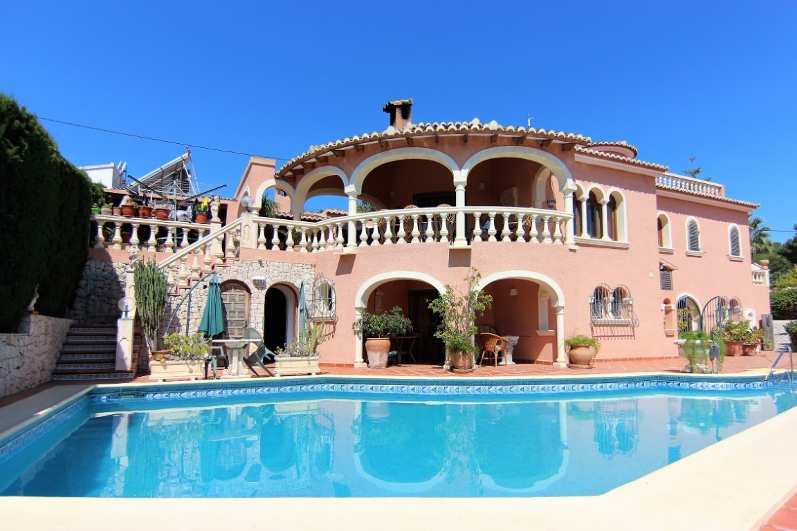 Vista exterior de la villa con piscina rectangular, destacando la fachada mediterránea en tonos salmón, amplias terrazas porticadas y balaustradas clásicas.