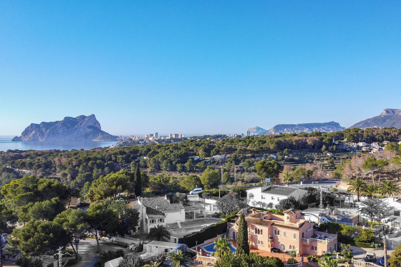 Vista panorámica aérea de la costa con el Peñón de Ifach al fondo, rodeada de abundante vegetación mediterránea y zonas residenciales bajo un cielo despejado.