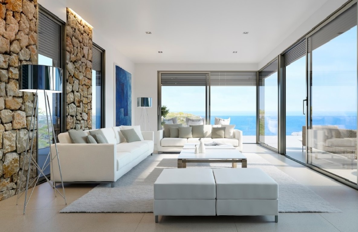 Salón luminoso con sofás blancos, pared de piedra natural con iluminación LED y grandes ventanales que ofrecen vistas panorámicas al mar Mediterráneo.