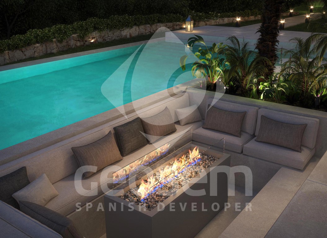 Salón exterior hundido junto a la piscina con chimenea de gas moderna, cojines grises y vegetación tropical iluminada bajo un muro de piedra natural.