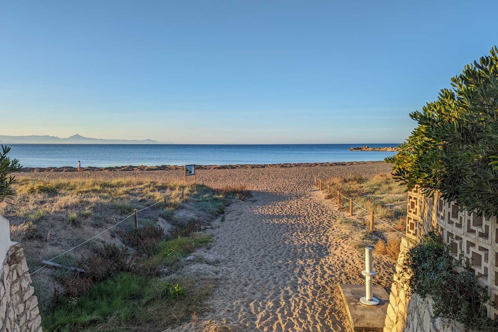 Acceso directo a la playa de arena a través de un sendero natural, con vistas panorámicas al mar Mediterráneo y las montañas en el horizonte.