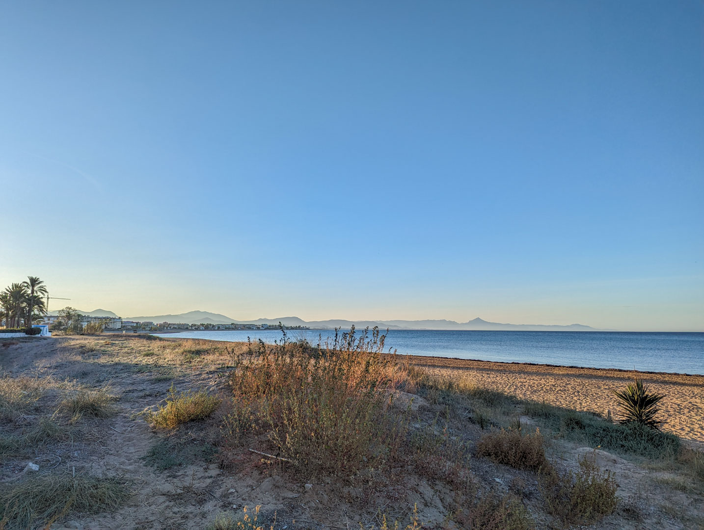 Vista de la playa natural próxima a la propiedad, con dunas de arena, vegetación autóctona y siluetas montañosas al fondo bajo un cielo despejado.