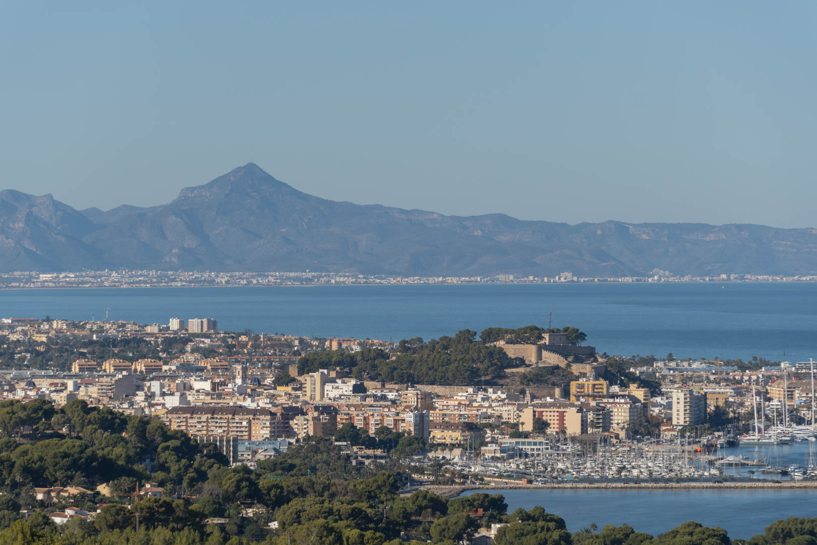 Vista panorámica elevada de Denia que muestra el castillo histórico, el puerto deportivo con sus embarcaciones y la silueta de las montañas sobre el mar Mediterráneo.