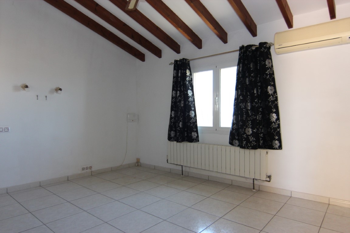 Habitación con techo inclinado y vigas de madera oscura, suelo de baldosas claras, radiador bajo la ventana y aire acondicionado tipo split.