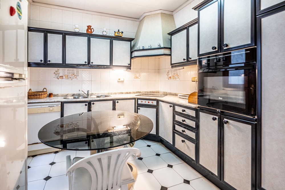 Cocina funcional con mobiliario de paneles blancos y marcos oscuros, equipada con electrodomésticos integrados, encimera de mármol y mesa de cristal sobre suelo cerámico.