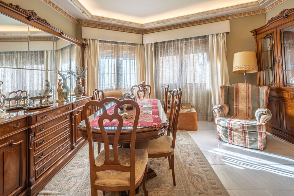 Comedor clásico con mobiliario de madera tallada, mesa con sobre de cristal, aparador con espejos y techo decorado con molduras e iluminación indirecta.