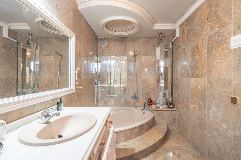 Cuarto de baño revestido íntegramente en mármol con bañera encastrada, lavabo sobre encimera blanca y un detalle decorativo circular en el techo.