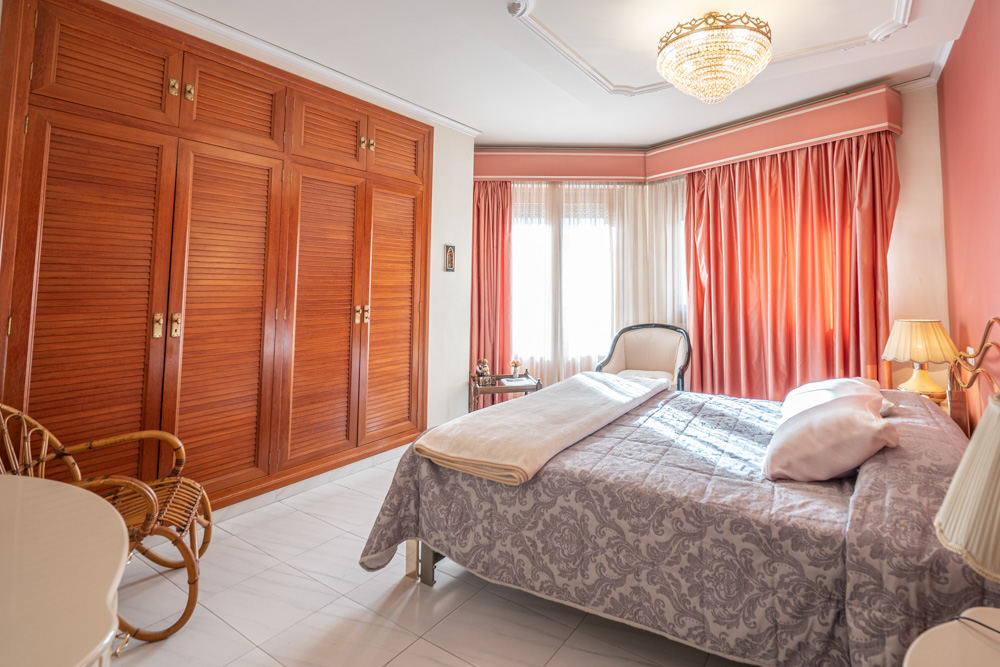 Dormitorio con armarios empotrados de madera maciza, ventanal con cortinas en tonos rosados, suelo cerámico y una elegante lámpara de araña de cristal.